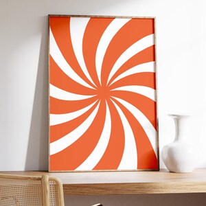 Op de afbeelding: Een ingelijst print met een swirlend oranje en wit patroon. De print hangt aan een witte muur boven een houten bureau met een stoel.
