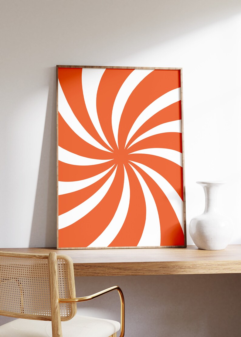 Op de afbeelding: Een ingelijst print met een swirlend oranje en wit patroon. De print hangt aan een witte muur boven een houten bureau met een stoel.