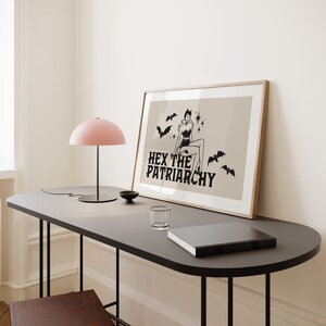 May include: A framed print featuring a black and white illustration of a woman with bat wings and a star on her forehead. The text "HEX THE PATRIARCHY" is beneath the illustration. The print is on a beige background and is framed in a light wood frame. The print is resting on a black table with a pink lamp and a glass on the table.
