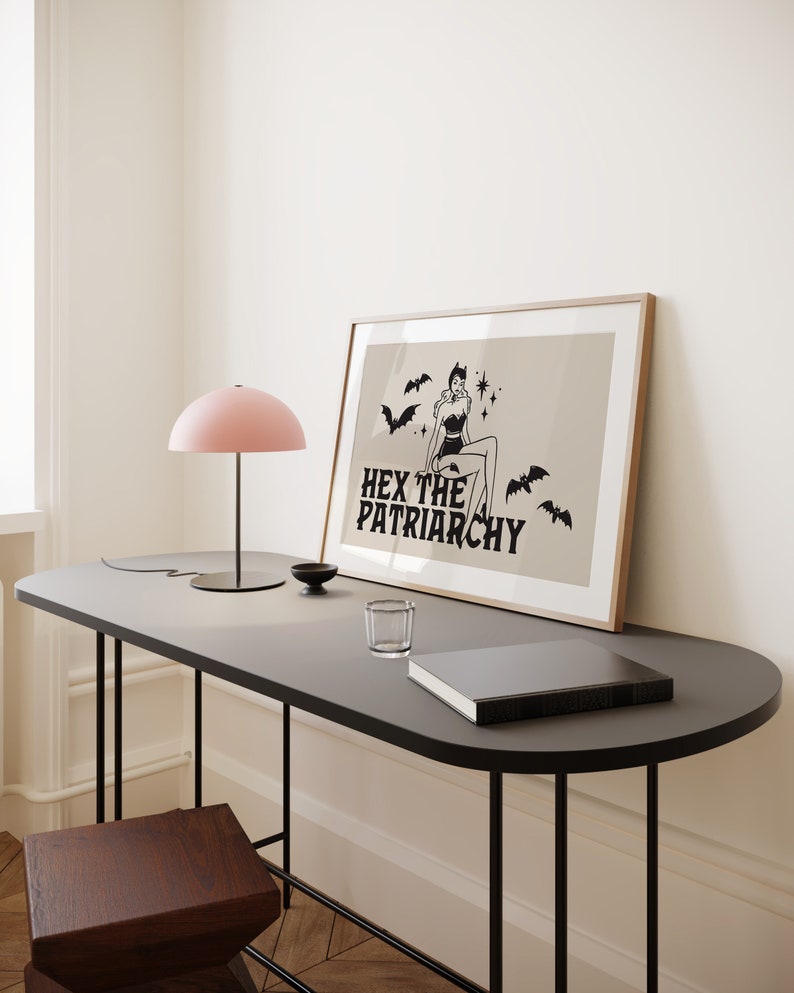 May include: A framed print featuring a black and white illustration of a woman with bat wings and a star on her forehead. The text "HEX THE PATRIARCHY" is beneath the illustration. The print is on a beige background and is framed in a light wood frame. The print is resting on a black table with a pink lamp and a glass on the table.