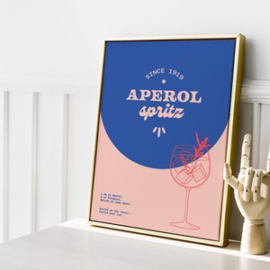 May include: A framed poster with a blue and pink background. The poster features a stylized illustration of a glass of Aperol Spritz with the text "Aperol Spritz" and "Since 1919". The poster also includes the recipe for Aperol Spritz.