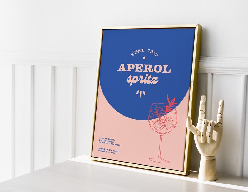 May include: A framed poster with a blue and pink background. The poster features a stylized illustration of a glass of Aperol Spritz with the text "Aperol Spritz" and "Since 1919". The poster also includes the recipe for Aperol Spritz.
