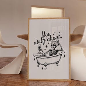 Funny Halloween decor. Light beige background with black foreground. Illustration of a skeleton sitting in a vintage bathtub with a glass of wine and text that reads you dirty ghoul. Perfect bathroom decor for the fall autumn season.