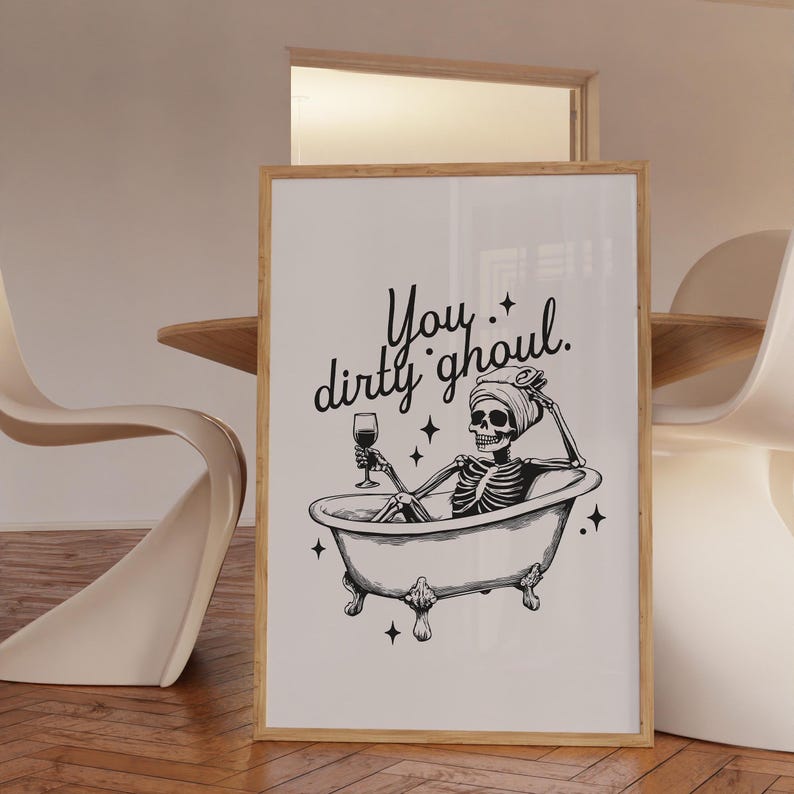 Funny Halloween decor. Light beige background with black foreground. Illustration of a skeleton sitting in a vintage bathtub with a glass of wine and text that reads you dirty ghoul. Perfect bathroom decor for the fall autumn season.