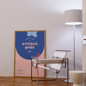 May include: A framed poster with a blue background and pink circle. The poster says "Since 1919 Aperol Spritz" and includes a recipe for the drink. A white chair with a metal frame sits in front of the poster.