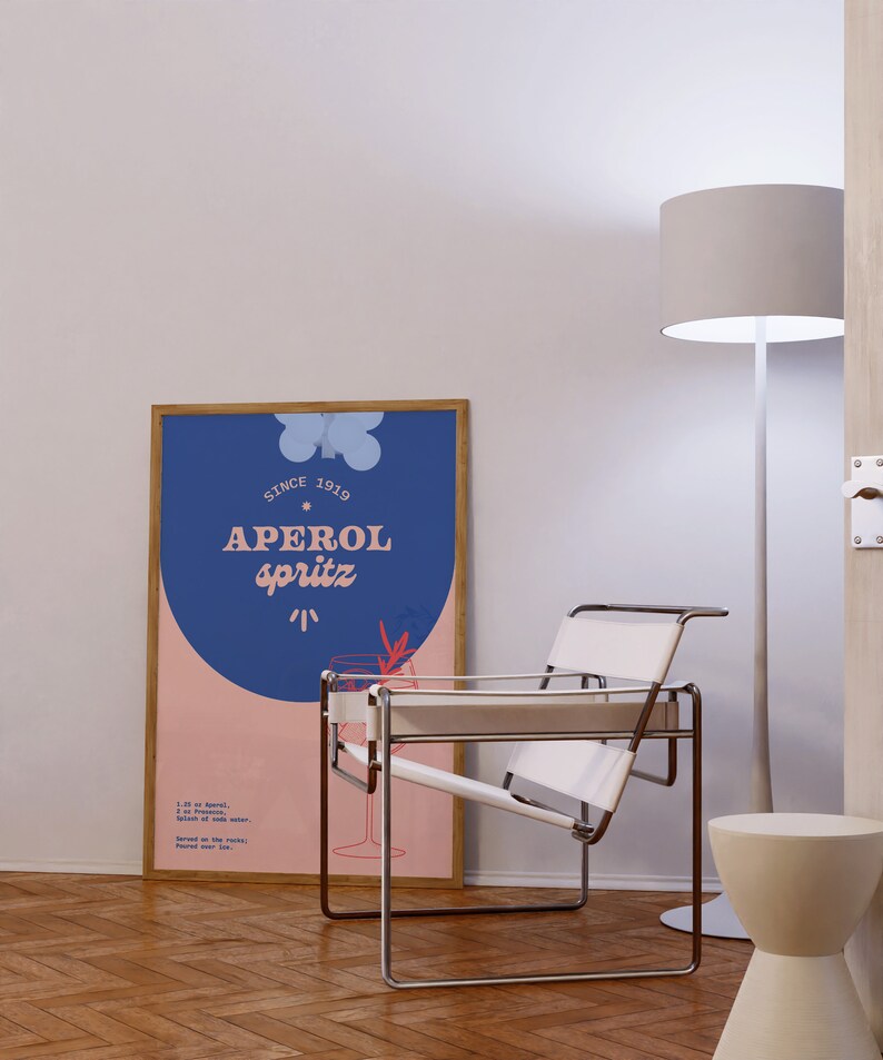 May include: A framed poster with a blue background and pink circle. The poster says "Since 1919 Aperol Spritz" and includes a recipe for the drink. A white chair with a metal frame sits in front of the poster.