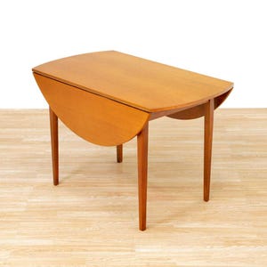 May include: A light brown, mid-century modern drop-leaf table. The table has a rectangular top with rounded corners and a drop-leaf on each side. The table is supported by four tapered legs. The table is approximately 30 inches long when the leaves are down.