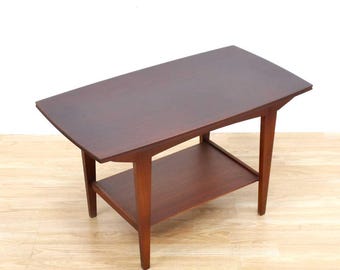May include: A mid-century modern style coffee table with a dark brown finish. The table has a rectangular top with rounded corners and a lower shelf. The legs are tapered and angled, and the table is made of wood.