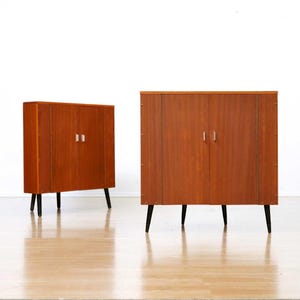 May include: Two mid-century modern wooden cabinets with a warm brown finish. Each cabinet has two doors with silver handles and black tapered legs. The cabinets are set against a white background.