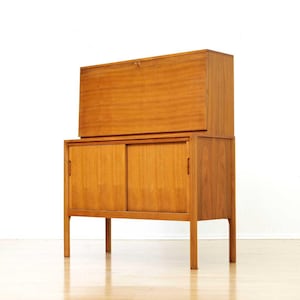 May include: A mid-century modern wooden cabinet with a light brown finish. The cabinet has a top section that opens upwards and a lower section with sliding doors. The cabinet stands on four slender legs.