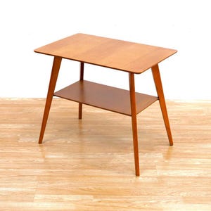 May include: A mid-century modern side table with a light wood finish and two shelves. The table has a rectangular top and angled legs.