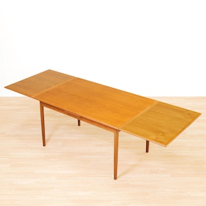 May include: A rectangular wooden dining table with a light brown finish and a smooth surface. The table features two extendable leaves on either side, supported by four tapered legs that angle outward from the center of the frame.