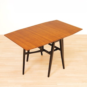 May include: A rectangular dining table featuring a warm, medium-brown wood grain tabletop with a visible seam down the center. The table is supported by four tapered, solid black legs connected by horizontal support bars in a matching black finish.