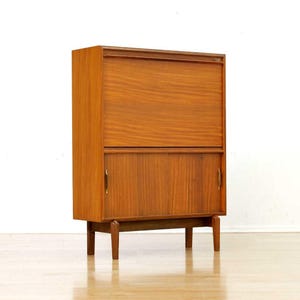 May include: A mid-century modern wooden cabinet with a warm brown finish. The cabinet features a top section with a drop-down door and a lower section with sliding doors. It stands on four tapered legs and has brass-colored handles.