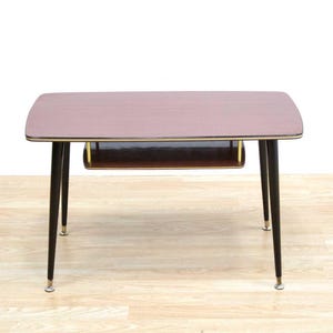 May include: A mid-century modern coffee table with a rectangular top and a lower shelf. The tabletop is a reddish-brown color with a gold trim. The legs are black with gold-colored tips. The table measures approximately 40 inches long, 20 inches wide, and 18 inches high.