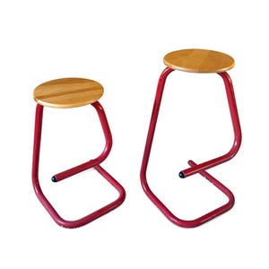 May include: Two stools with a minimalist design. Each stool has a round, light-colored wooden seat and a red metal frame. The frames have a unique, curved shape. The smaller stool is approximately 18 inches tall, and the larger one is about 30 inches tall.