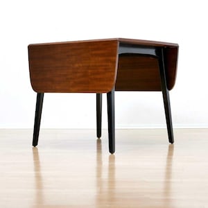 May include: A drop-leaf table featuring a rectangular top with a rich brown wood grain finish and two hinged side panels. The table is supported by four straight, solid black legs that provide a stable base for the wooden surface structure.