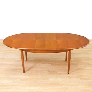 May include: An oval wooden dining table with a warm brown finish and a smooth surface. The table features four tapered legs that extend outward from the center, supporting the tabletop which shows a visible seam indicating an extendable design.