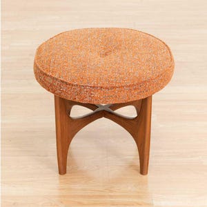 May include: An orange and brown mid-century modern stool. The round seat is upholstered in a textured orange fabric with a button in the center. The wooden legs are a warm brown color and have a unique, curved design.