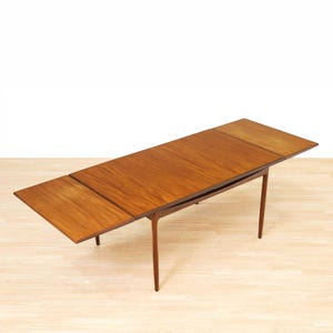May include: A rectangular wooden dining table with a warm brown finish and visible wood grain. The table features an extendable design with a central main surface and two side leaves that are partially extended, supported by four tapered legs.