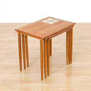 May include: A set of three nesting tables with a light brown wood finish. The tables have rectangular tops and slender, tapered legs. The top of the largest table features a decorative tile inlay with a geometric pattern. The tables are designed to stack for space-saving storage.