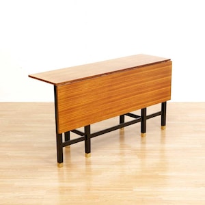 May include: A mid-century modern drop-leaf table with a rectangular top and a warm wood grain finish. The table has black legs with gold-colored accents on the feet. The table is partially open, with one leaf extended.