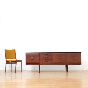 May include: A long wooden sideboard featuring three cabinets with rectangular panels and central handles, alongside a central column of three drawers. Beside it stands a wooden chair with a mustard yellow upholstered back and seat.