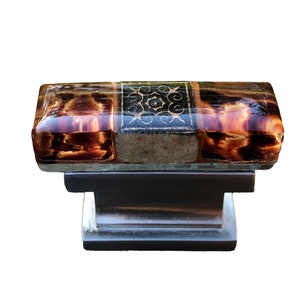 May include: A decorative cabinet knob with a rectangular, brown, and amber-colored top. The top features a dark, ornate design in the center. The knob sits on a dark, rectangular base with a silver accent. This hardware is suitable for furniture.