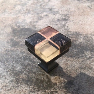 May include: Square cabinet knob with a black base and a multi-colored glass top. The glass top is made of four squares, two of which are black, one is light brown, and one is light yellow.
