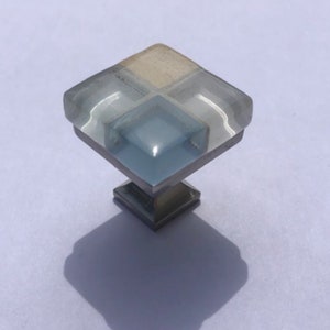 May include: Square cabinet knob with a clear glass base and a blue and white geometric design. The knob has a silver metal base.