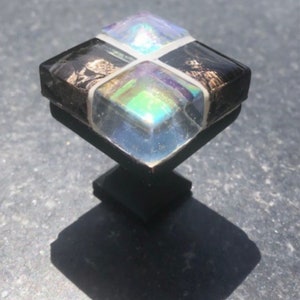 May include: Square cabinet knob with a black base and a multi-colored iridescent glass top. The glass top is divided into four sections, each with a different color.
