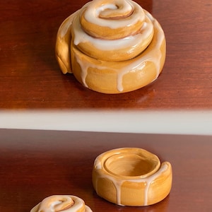 Puede incluir: Dos recipientes de cerámica con forma de rollo de canela con glaseado blanco. El recipiente más grande está cerrado y el más pequeño está abierto.