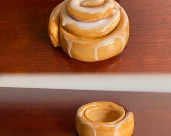 May include: Two ceramic cinnamon roll shaped containers with white icing. The larger container is stacked on top of itself. The smaller container is sitting on a wooden surface.