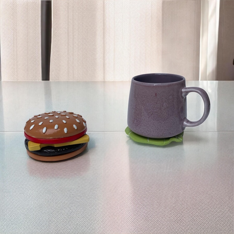 May include: A purple ceramic mug with a handle sits on a green leaf-shaped coaster. A toy hamburger with sesame seeds and a yellow cheese slice is on a white surface.