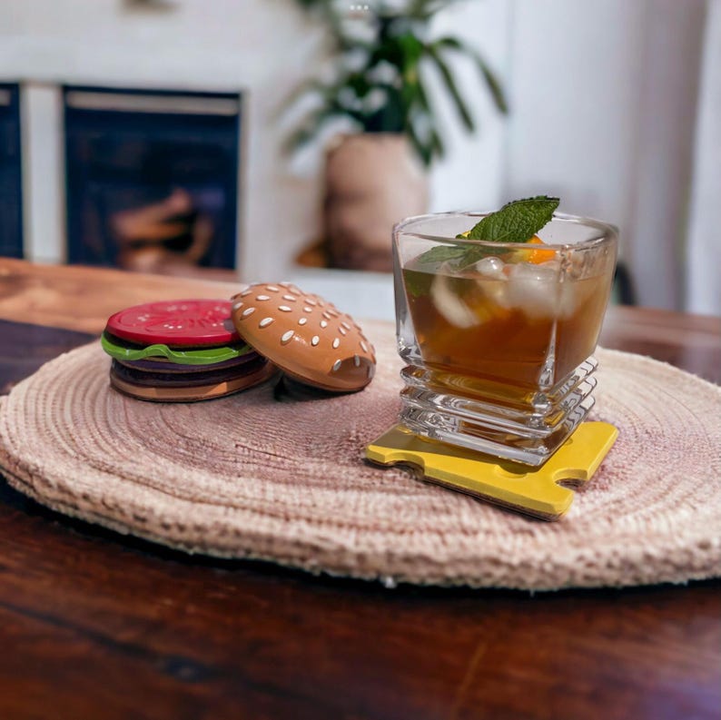 May include: Two toy hamburger patties on a wooden coaster with a glass of iced tea on a yellow cheese-shaped coaster.