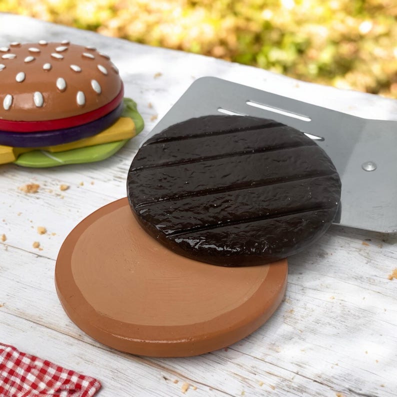 May include: Two brown and black toy hamburger patties, one with grill marks, on a white wooden surface. A toy spatula and a toy hamburger bun are also visible.
