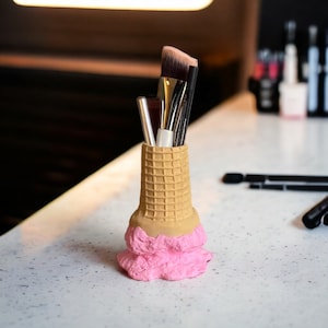 May include: A pink and brown ice cream cone shaped makeup brush holder with multiple brushes inside.