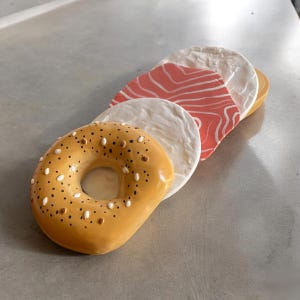 Op de afbeelding: Een stapel van vier ronde, keramische onderzetters in de vorm van voedsel. De bovenste onderzetter is een gele bagel met bruine en witte sprinkels. De tweede onderzetter is wit met een licht verhoogde rand. De derde onderzetter is rood met witte strepen. De onderste onderzetter is wit met een licht verhoogde rand.