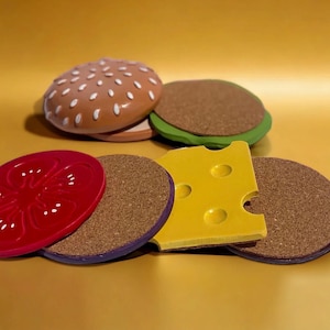 May include: Set of five colorful cork coasters shaped like a hamburger, tomato, cheese, and two burger buns. The coasters are made of cork and have a decorative design.