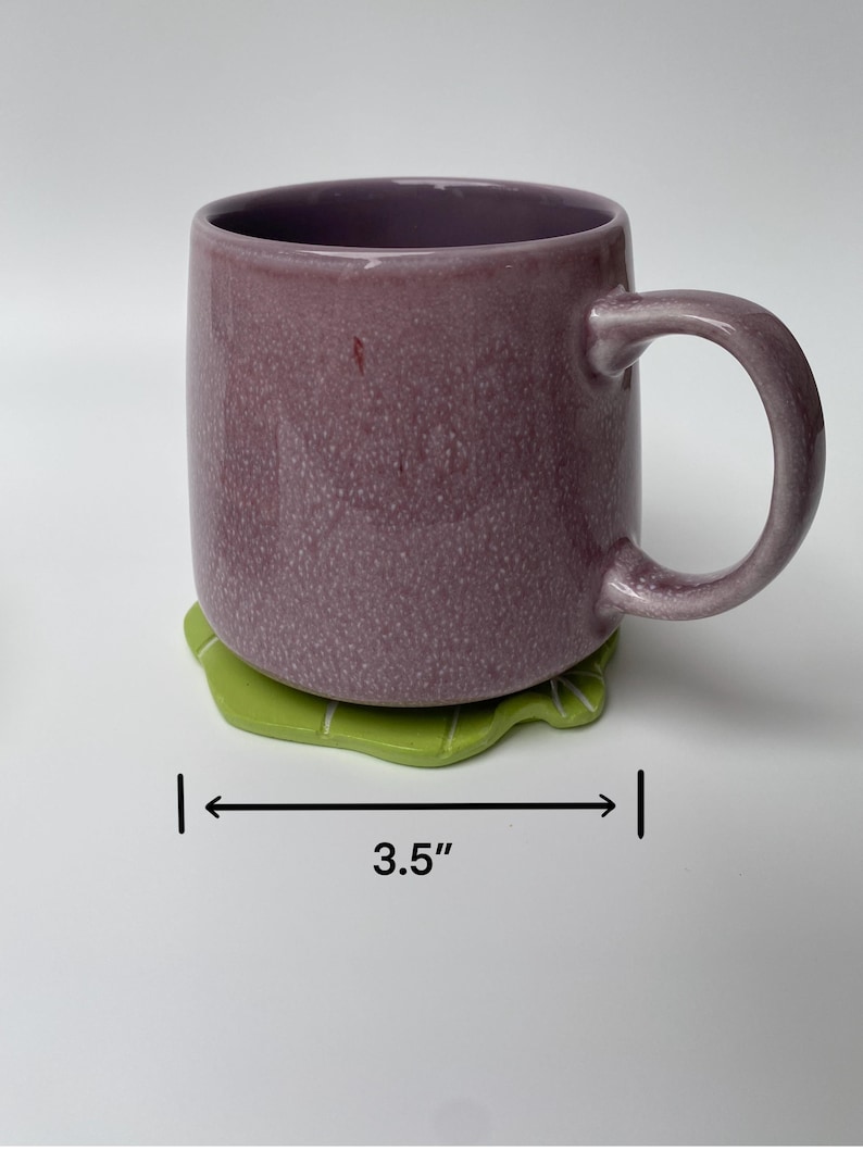 May include: A purple ceramic mug with a white interior and a green leaf-shaped coaster. The coaster is 3.5 inches wide.