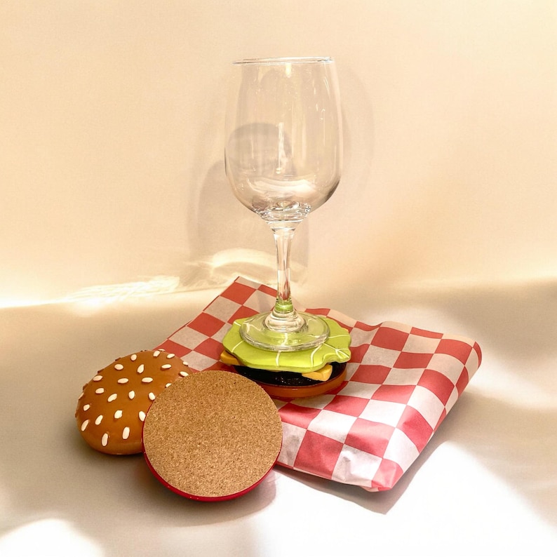 May include: A clear wine glass sits on a burger-shaped coaster. The coaster is on a red and white checkered paper. There is a second burger-shaped coaster next to the glass.