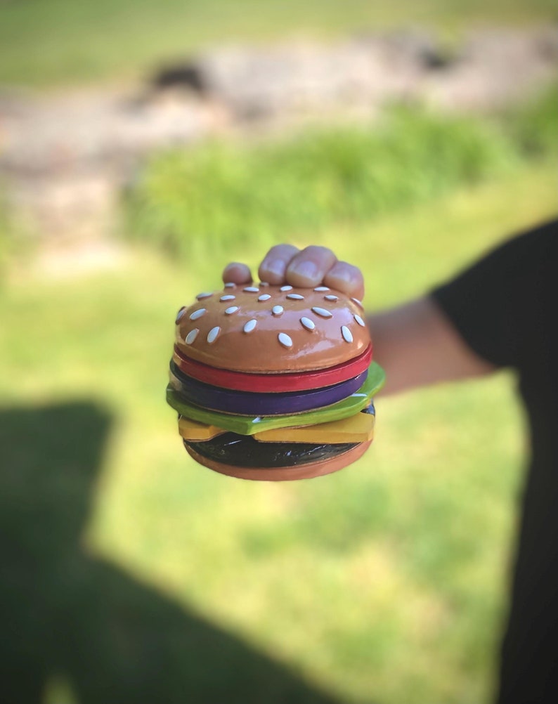 May include: A miniature wooden hamburger with a sesame seed bun, red tomato, green lettuce, purple onion, and yellow cheese.