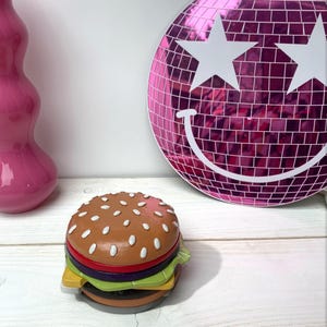 May include: A colorful, ceramic hamburger figurine with a white sesame seed bun, red tomato, green lettuce, and purple onion. The hamburger is sitting on a white wooden surface.