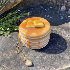 May include: A stack of ceramic pancakes with yellow icing and a gold chain necklace with a heart pendant.