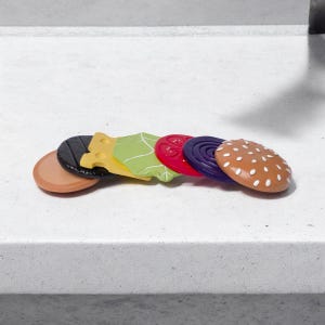 May include: A set of six wooden toy burger pieces, including a brown bun, a black sesame seed bun, a yellow cheese slice, a green lettuce leaf, a red tomato slice, and a purple onion slice.