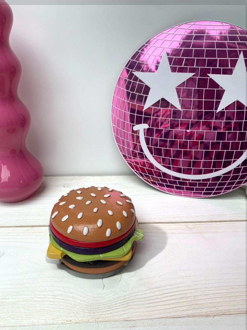 May include: A ceramic hamburger money box with a brown bun, red tomato, green lettuce, and yellow cheese. The money box is sitting on a white wooden surface in front of a pink disco ball with white stars.