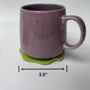 May include: A purple ceramic mug with a white interior and a green leaf-shaped coaster. The coaster is 3.5 inches wide.
