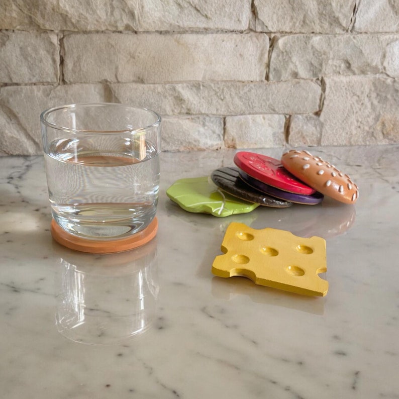 May include: A set of six colorful coasters shaped like different parts of a hamburger. The coasters are made of ceramic and feature a green lettuce leaf, a brown burger patty, a red tomato slice, a yellow cheese slice, a brown bun, and a white sesame seed bun. The coasters are arranged in a stack on a white marble surface. A glass of water sits on a brown coaster.