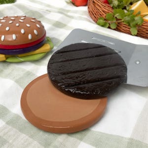 May include: A set of toy food items for pretend play, including a hamburger, a grilled patty, and a spatula. The hamburger is made of plastic and has a white bun, a red patty, and green lettuce. The grilled patty is brown and has grill marks. The spatula is silver and has a black handle.