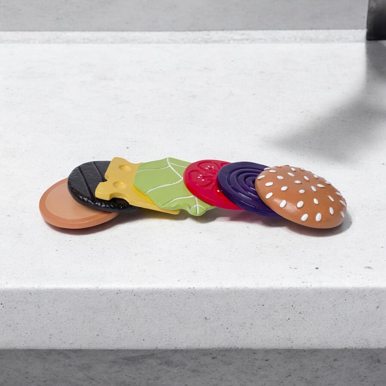 May include: A set of six wooden toy burger pieces, including a brown bun, a black sesame seed bun, a yellow cheese slice, a green lettuce leaf, a red tomato slice, and a purple onion slice.